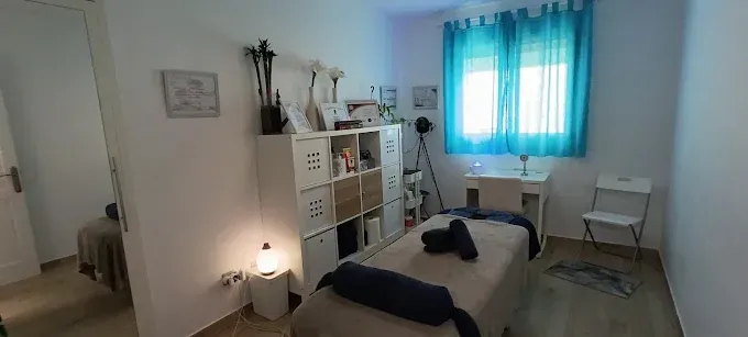 Una sala de terapia de masajes con una mesa de masajes, estantes de almacenamiento y una ventana con cortinas turquesas.