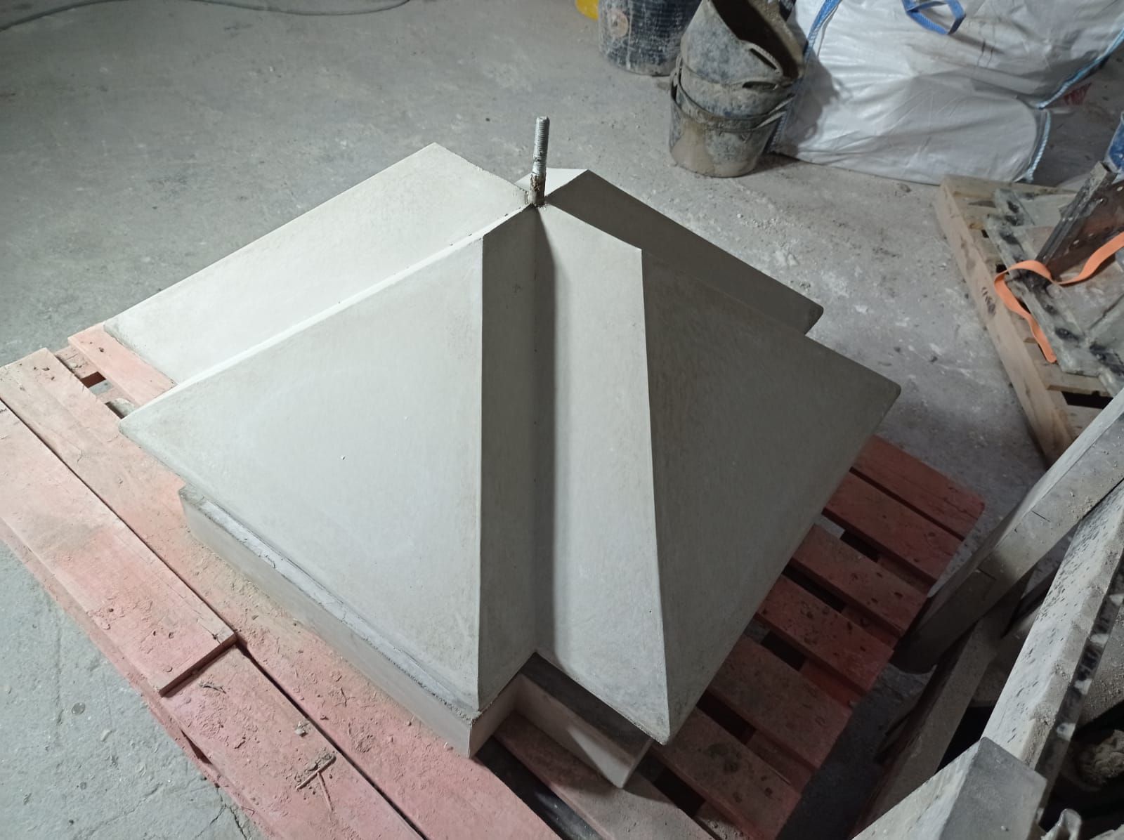 Estructura de hormigón gris y angular sobre un palé de madera, con una varilla metálica que se extiende hacia arriba.