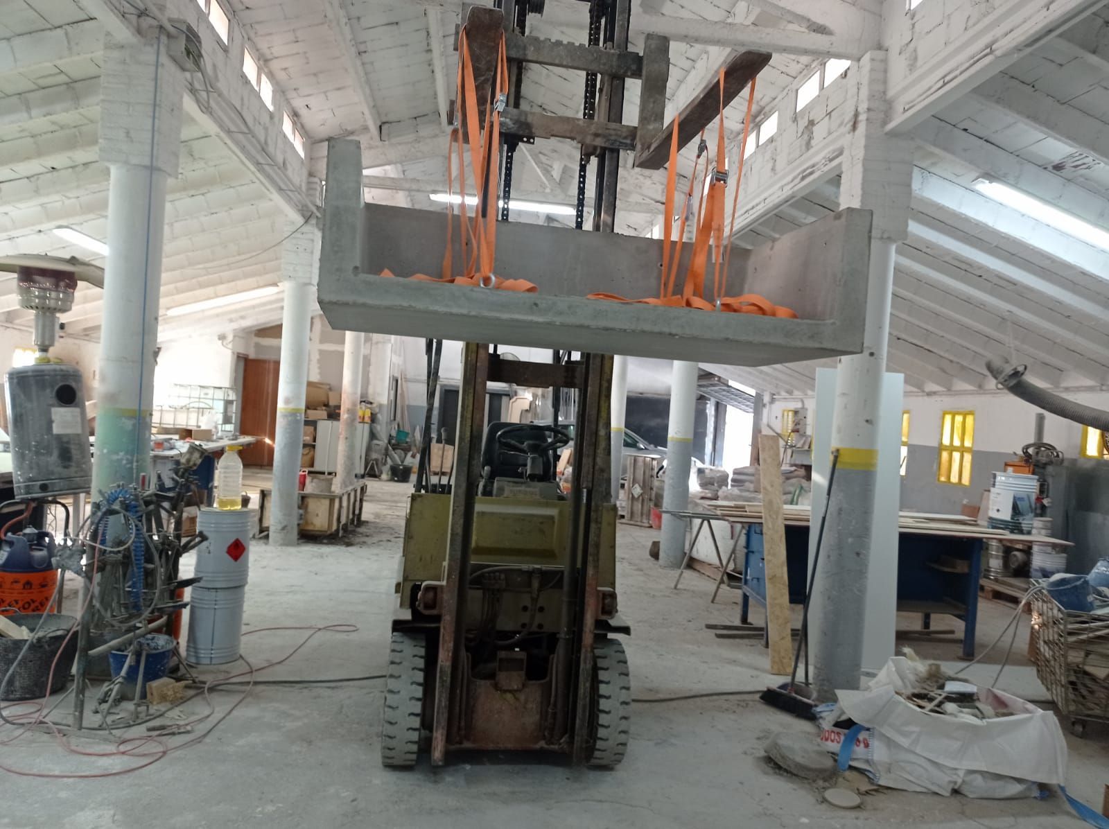 Una carretilla elevadora levanta un encofrado de hormigón dentro de un taller. Unas correas naranjas sujetan la carga.