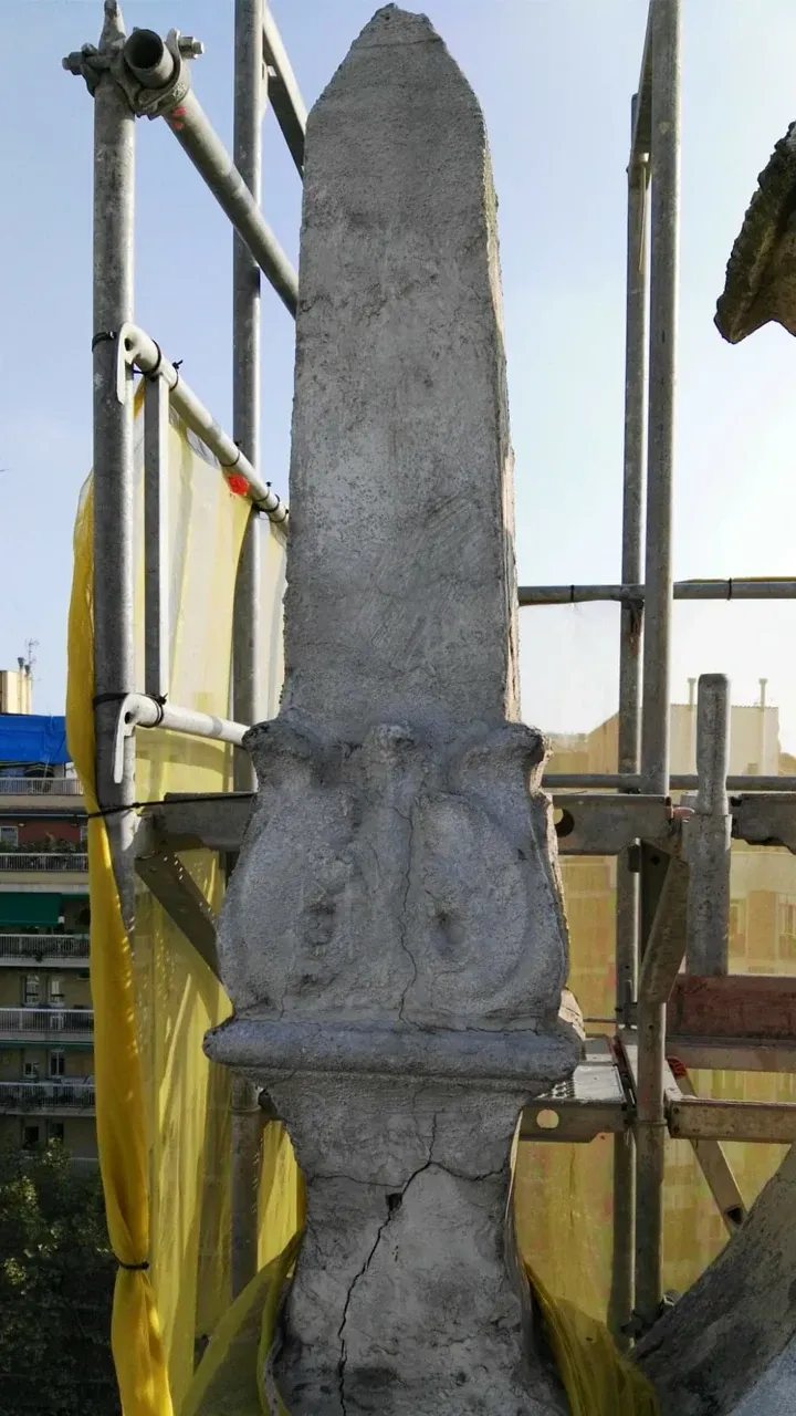 Alto obelisco de piedra erosionada, posiblemente arquitectónico, con tallas ornamentadas. Andamios y ciudad al fondo.
