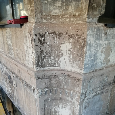 Esquina de un edificio con pintura descascarada. Superficies grises y blancas, con grietas y daños visibles.