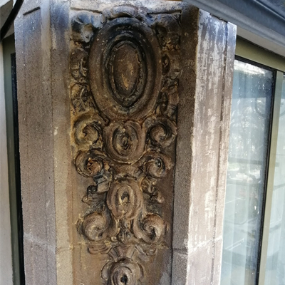 Columna de piedra desgastada y ornamentada con diseño decorativo ovalado y con volutas junto a una ventana.