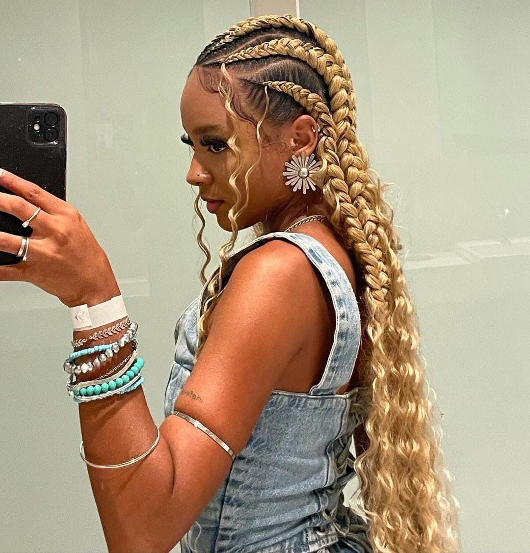 Mujer con trenzas rubias, vistiendo un top de mezclilla y joyas, tomándose una selfie frente al espejo.