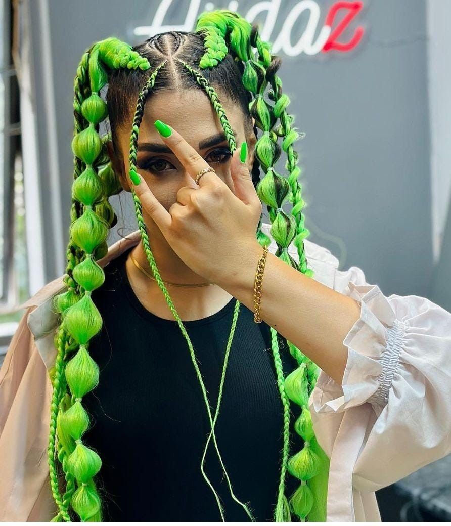 Mujer con trenzas de burbujas de color verde brillante, con top negro y levantando la mano.