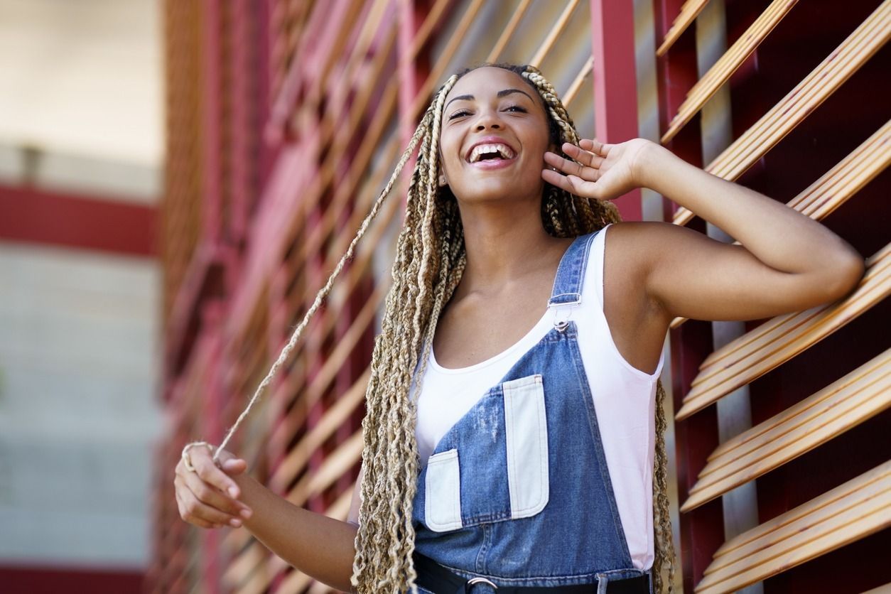 Mujer con mono y trenzas largas sonriendo, apoyada contra una pared roja y madera.