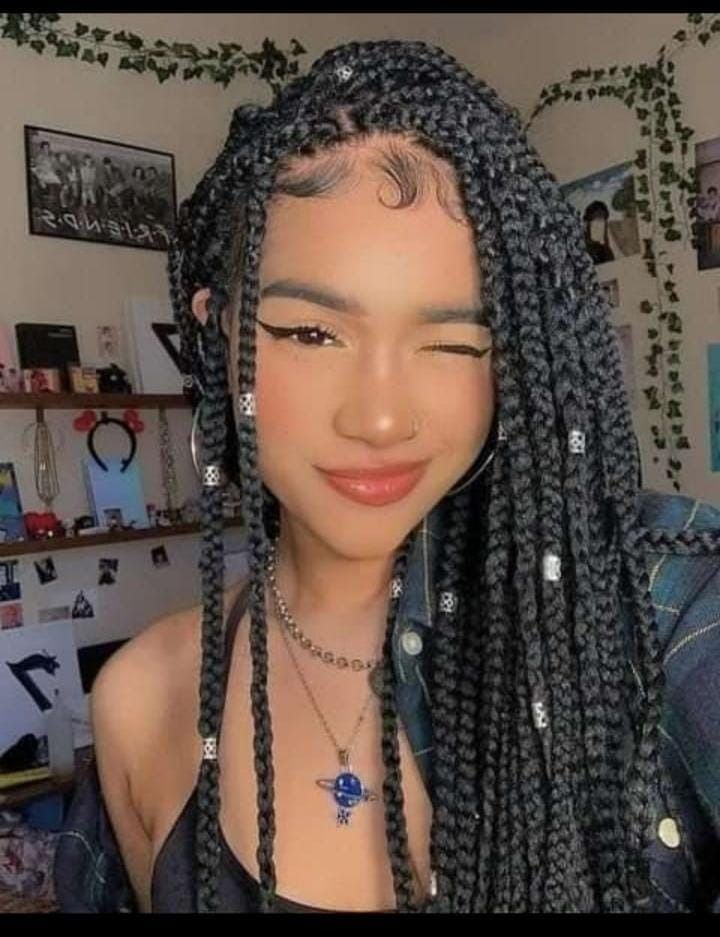Mujer joven con trenzas guiñando un ojo, luciendo un collar, sonriendo en una habitación con decoraciones.