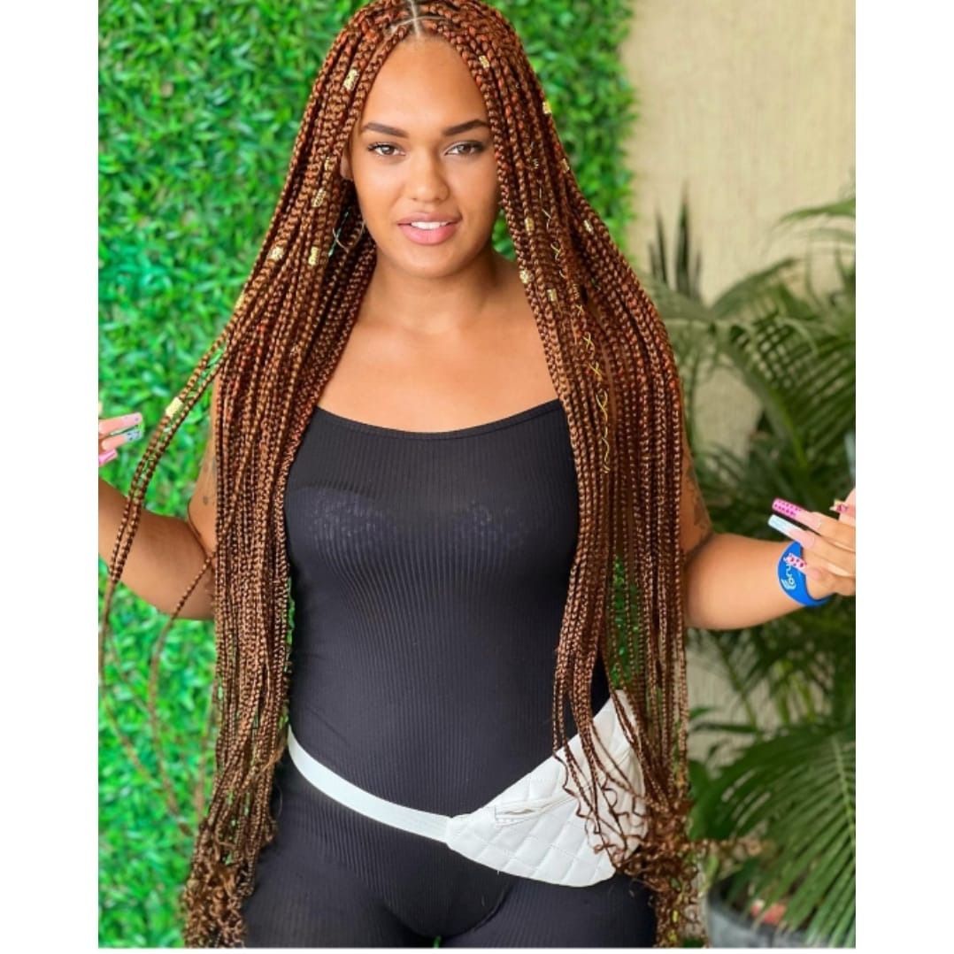 Mujer con largas trenzas marrones con un traje negro y una riñonera blanca, posando frente a la vegetación.