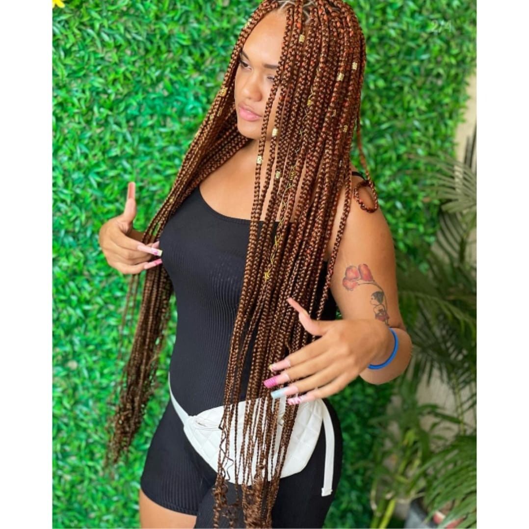 Mujer con largas trenzas castañas, vestida con un mono negro y una riñonera, posa frente a una pared verde.