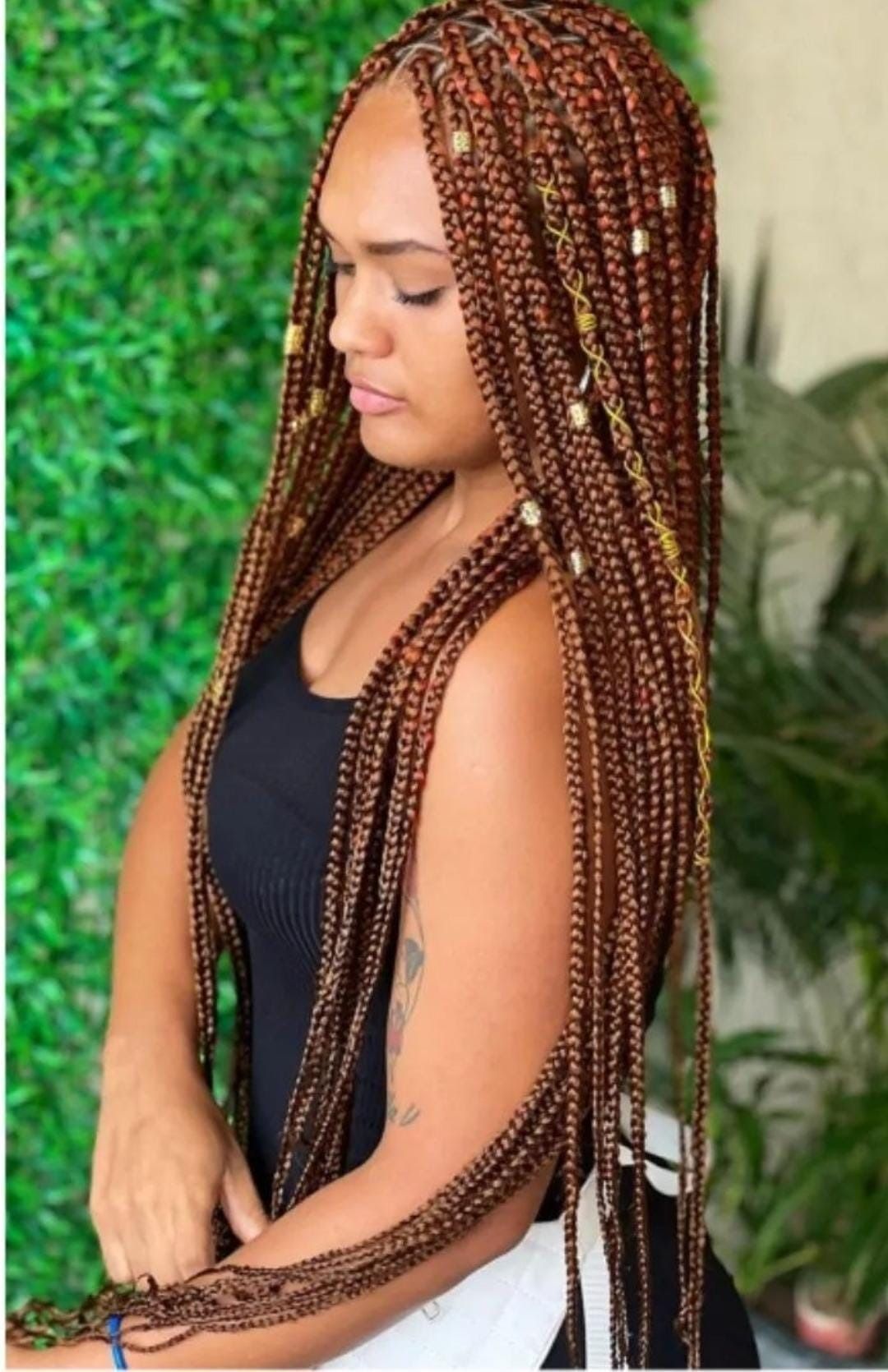 Mujer con largas trenzas de color cobre, parada frente a una pared verde, vistiendo una camiseta sin mangas negra.