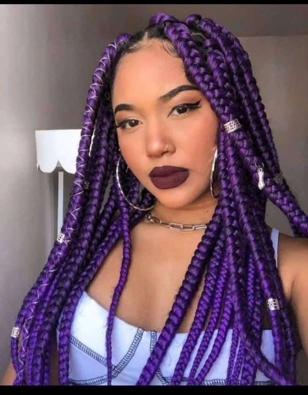 Mujer con trenzas de caja moradas, pendientes de aro, lápiz labial oscuro y top blanco.