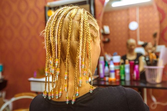 Cabello rubio en trenzas con cuentas, visto desde atrás en un salón con espejo y productos en un estante.