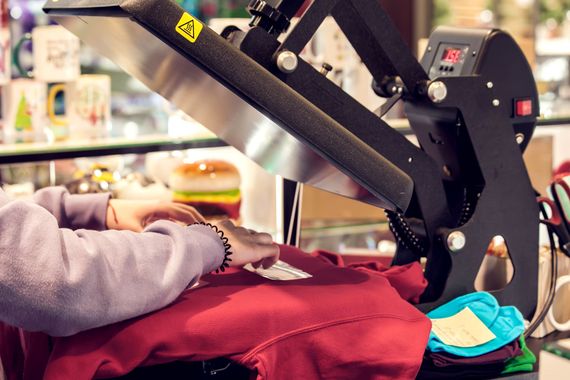 Eine Person bringt unter einer grossen, industriellen Transferpresse ein Design auf einen roten Kapuzenpullover auf.