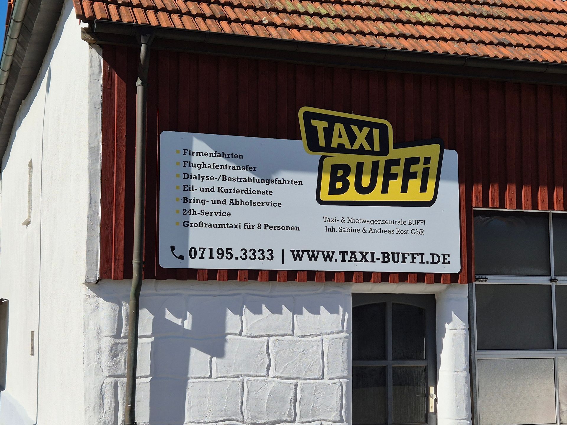 Ein Taxi-BUFFI-Schild an einem Gebäude, mit gelber und schwarzer Schrift und einer Telefonnummer/Webadresse darunter.