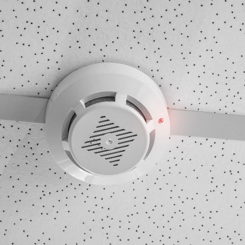 Détecteur de fumée fixé au plafond avec un voyant rouge, sur un plafond moucheté.