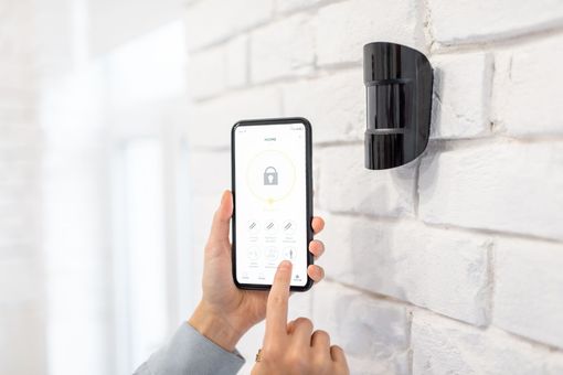 Des mains utilisent un smartphone pour contrôler une serrure connectée noire fixée au mur, sur un mur de briques blanches.