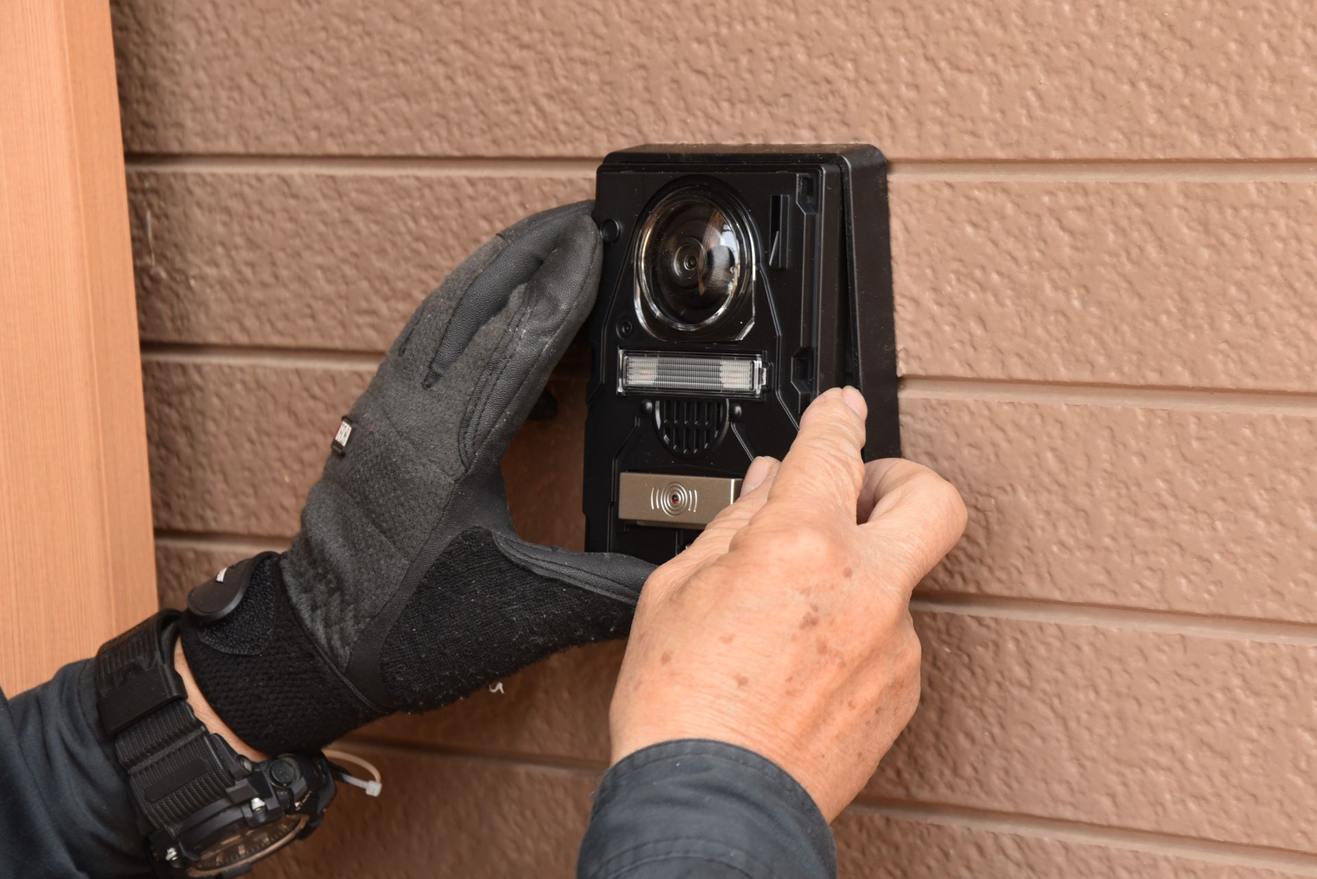 Des mains gantées installent une sonnette vidéo noire fixée au mur d'un mur extérieur marron.