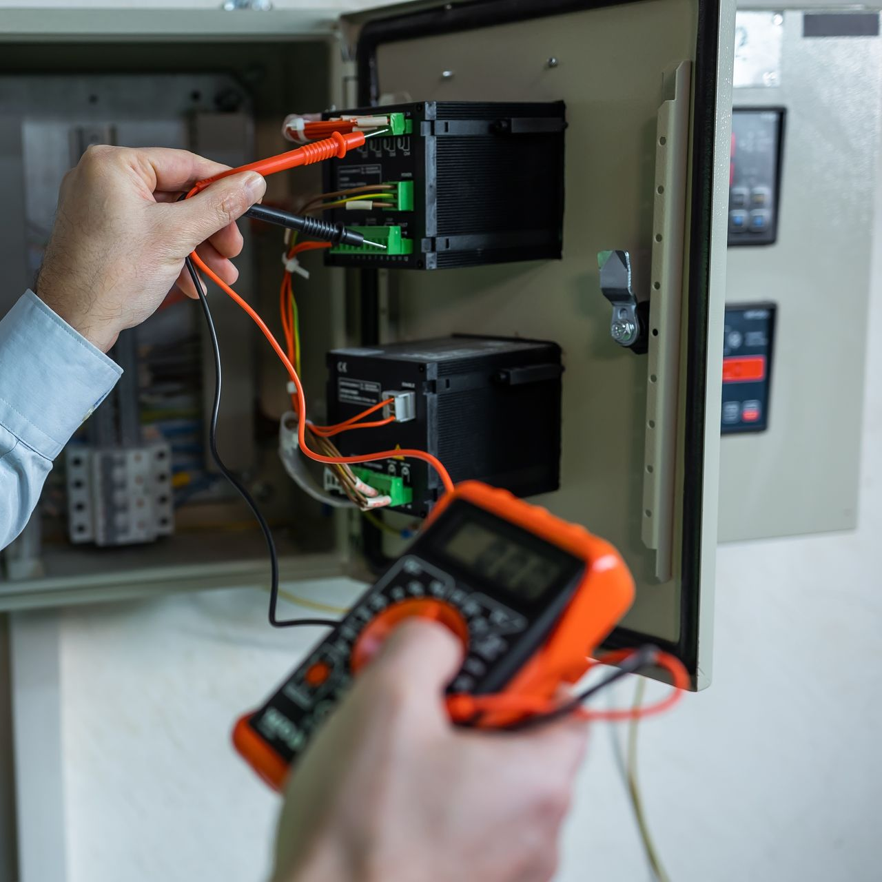 Un technicien teste un équipement électrique dans une armoire ouverte à l'aide d'un multimètre numérique et de sondes orange.
