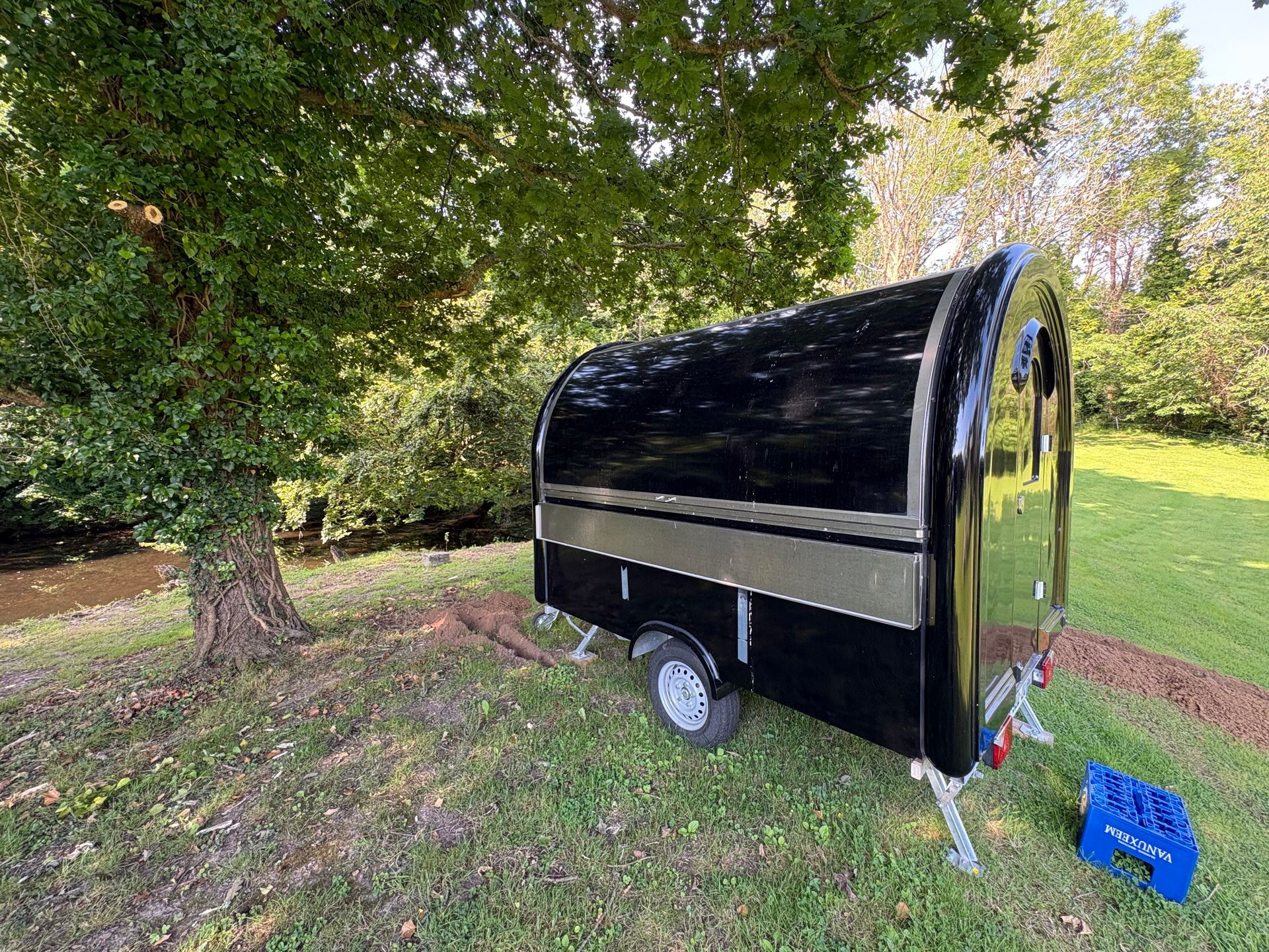 Une caravane noire est garée sur l'herbe sous des arbres, avec une poubelle bleue à proximité.