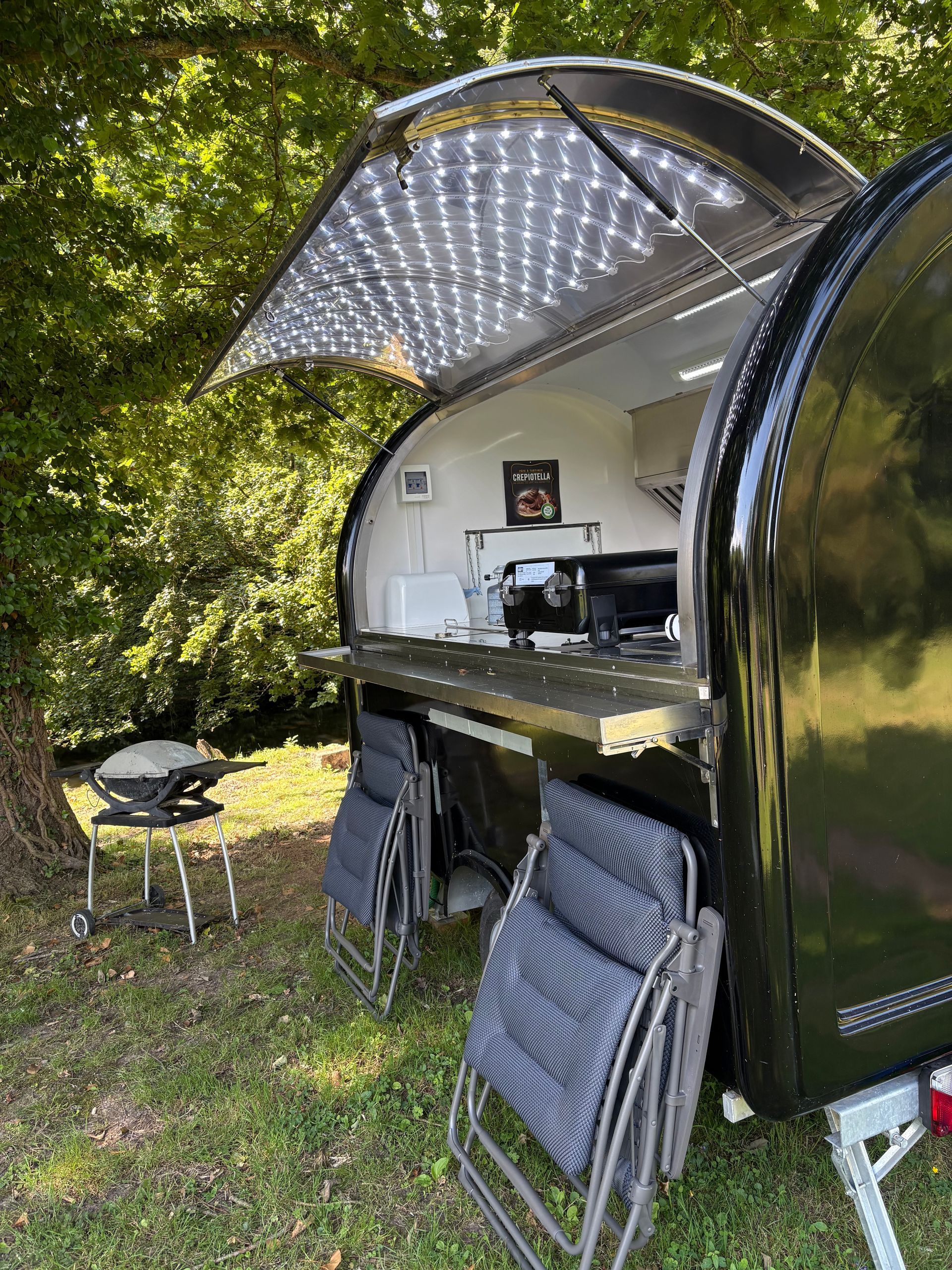 Caravane pliante ouverte avec auvent, cuisine extérieure et chaises pliantes dans un camping arboré.