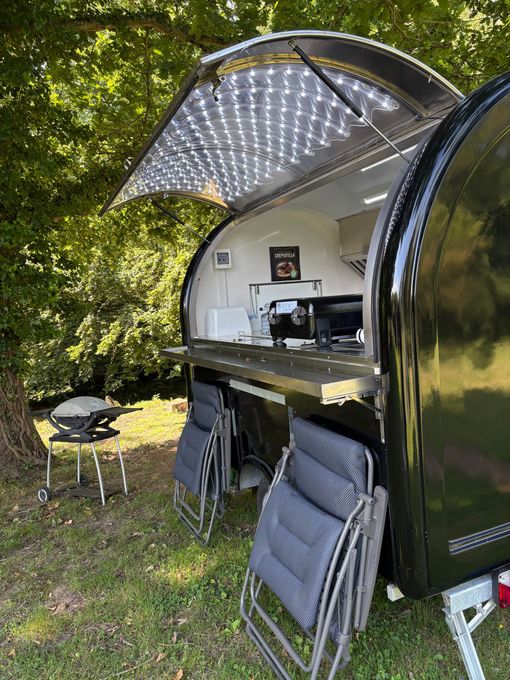 Caravane pliante ouverte avec auvent, cuisine extérieure et chaises pliantes dans un camping arboré.