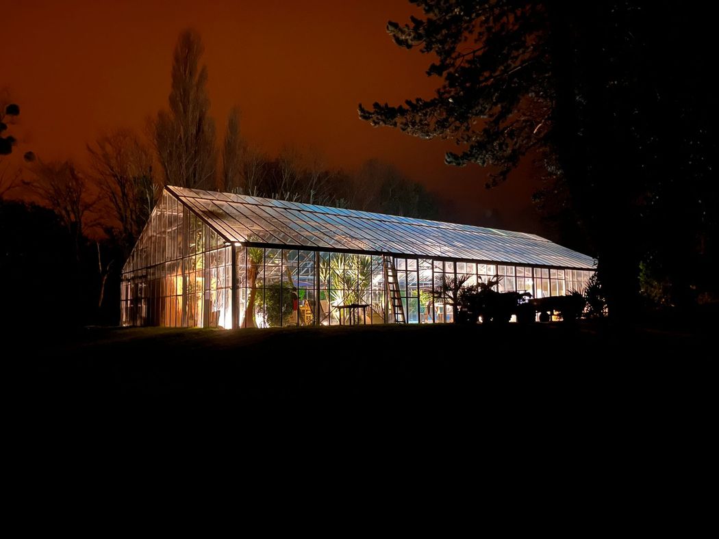 Serre illuminée la nuit, avec des lumières chaudes qui contrastent avec un ciel orange foncé.