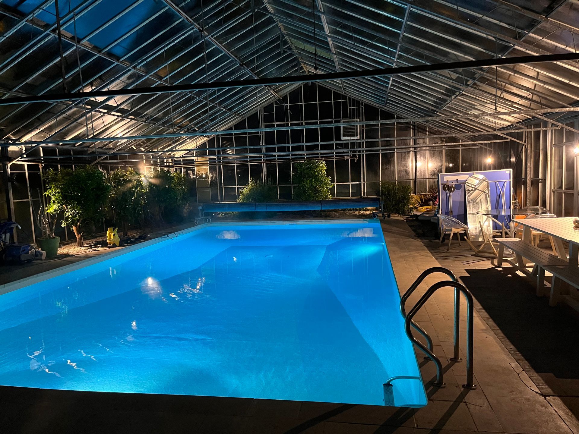 Piscine intérieure ouverte la nuit, avec une eau bleue sous un toit de verre, des chaises longues et des lumières.