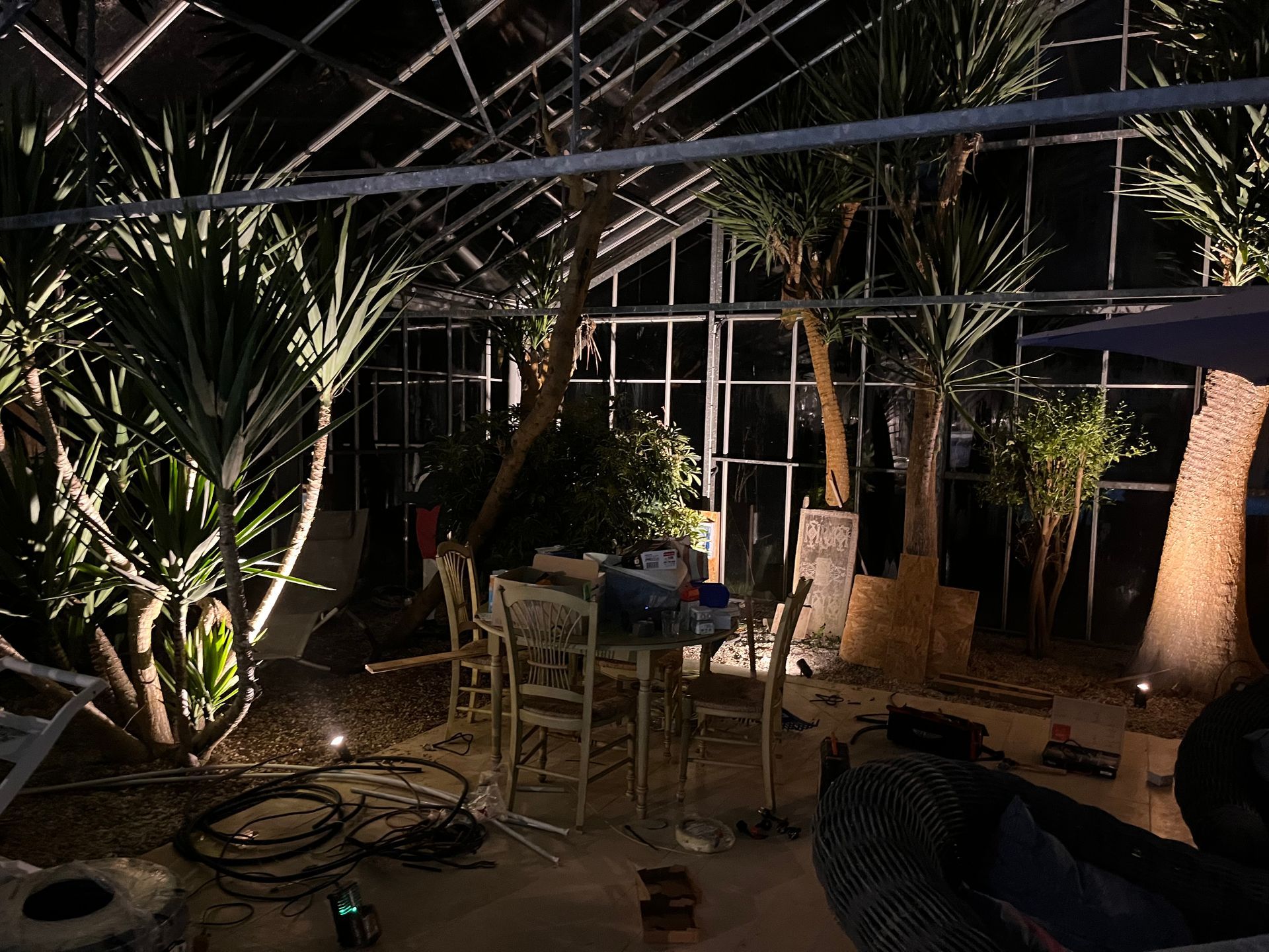 Patio extérieur faiblement éclairé avec de grandes plantes, des guirlandes lumineuses, une table et des chaises la nuit.