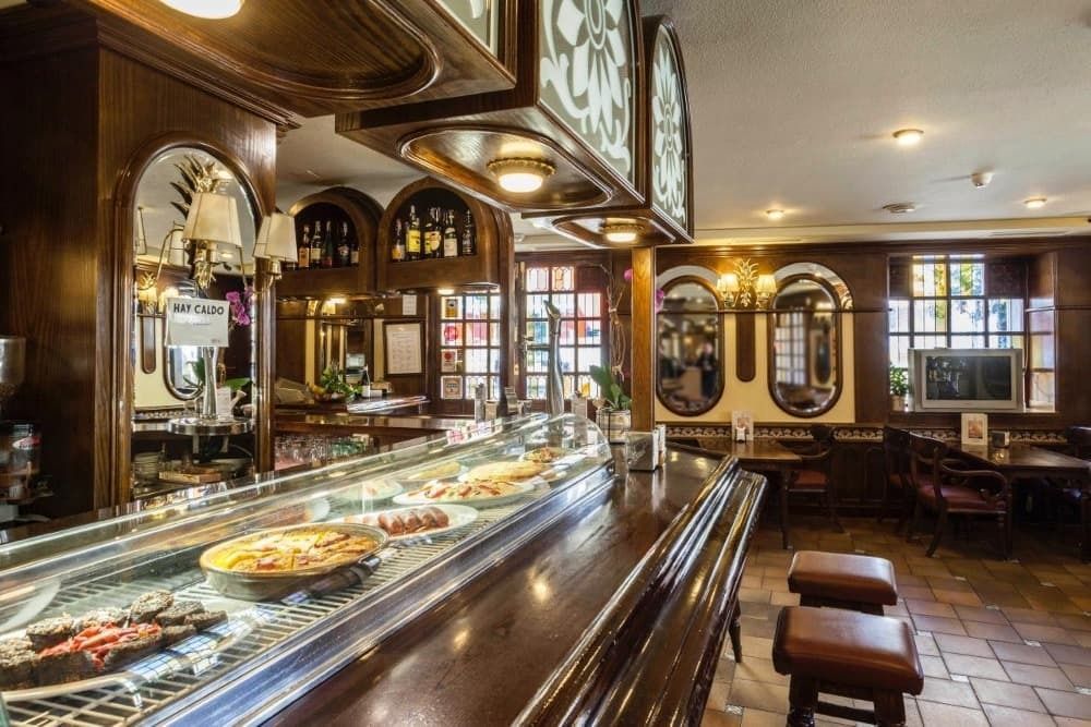 Interior de bar con un largo mostrador con comida. Revestimiento de madera oscura y espejos.