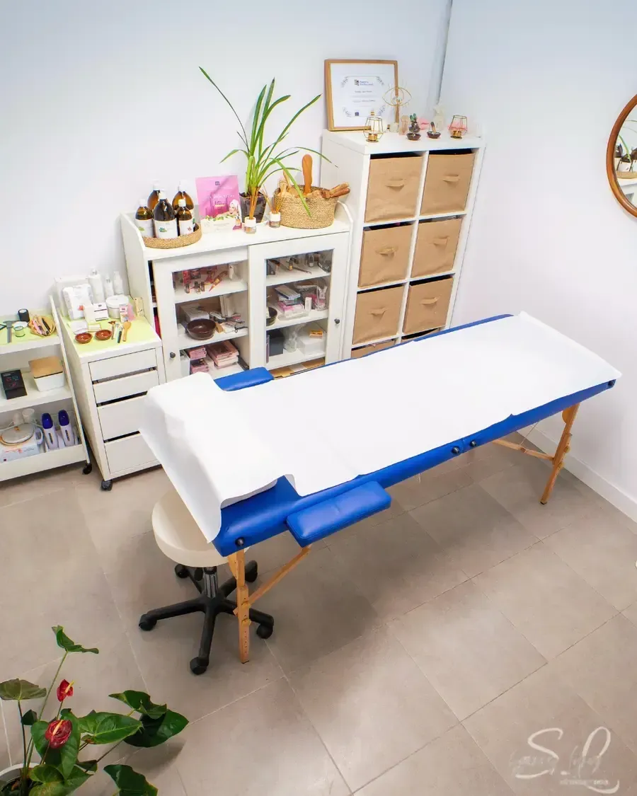 Camilla de masajes en habitación luminosa con almacenaje, plantas y taburete.