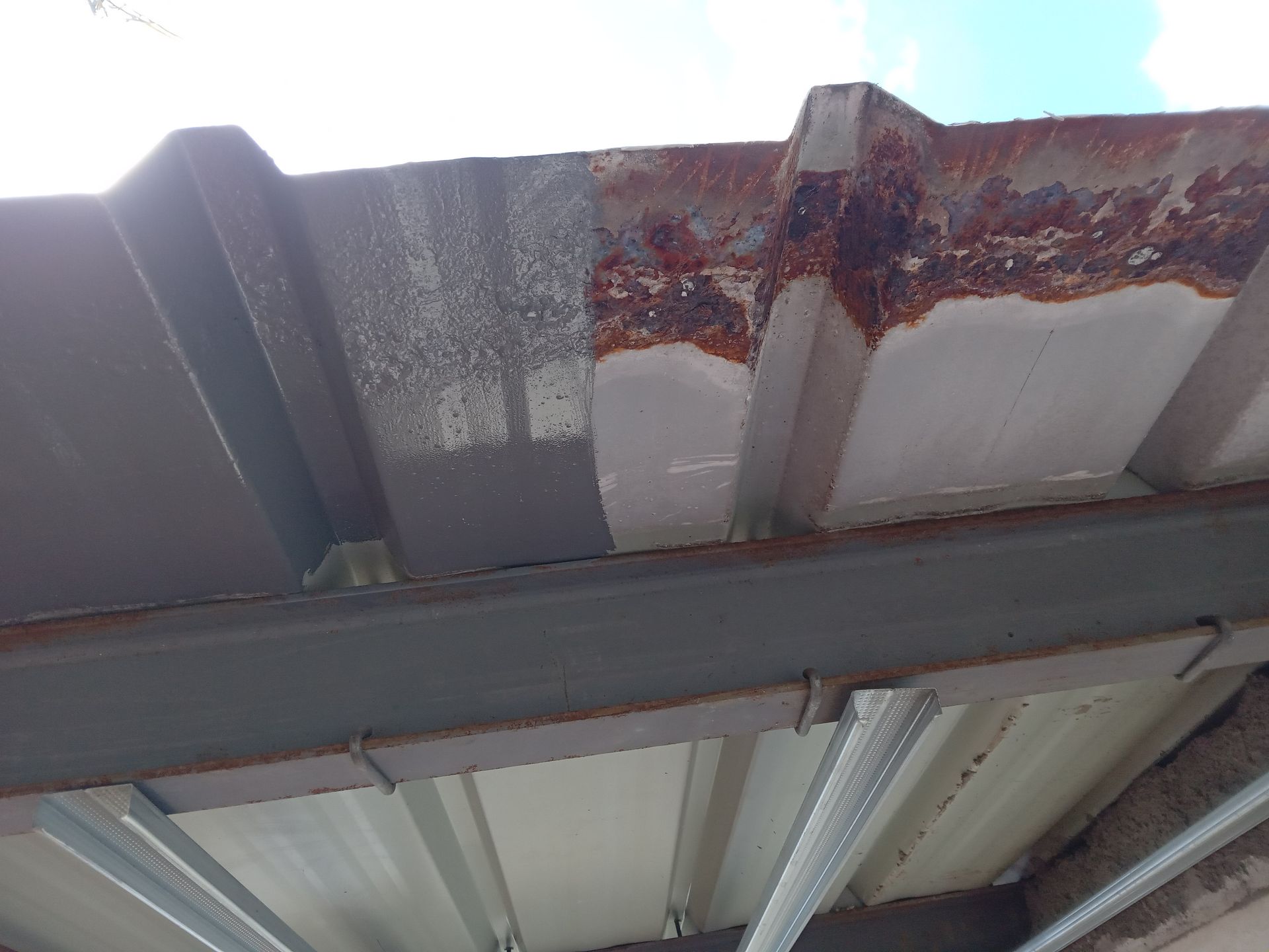 Bordure de toit en métal rouillé, peinte en gris et blanc, soutenue par une poutre et une charpente métalliques grises.