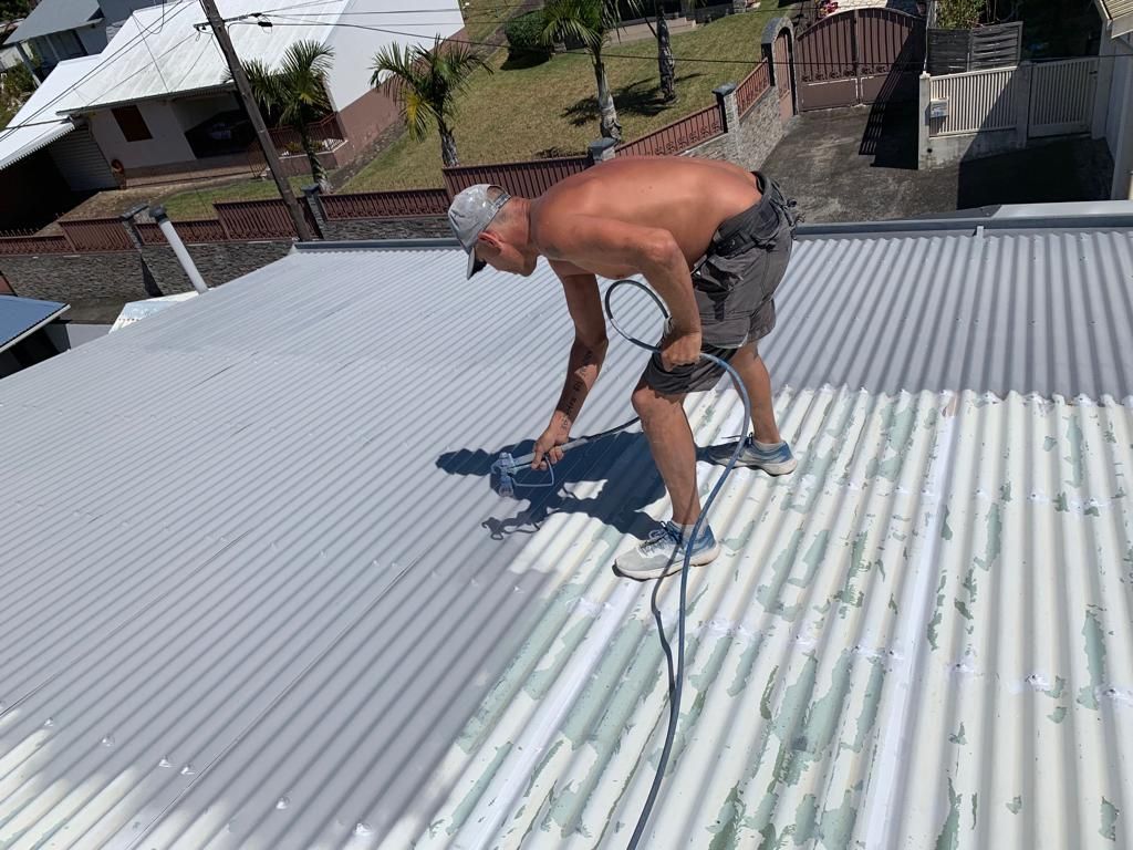 Un homme peint un toit en tôle ondulée. Torse nu, vêtu d'un short et d'une casquette, il travaille dehors par une journée ensoleillée.