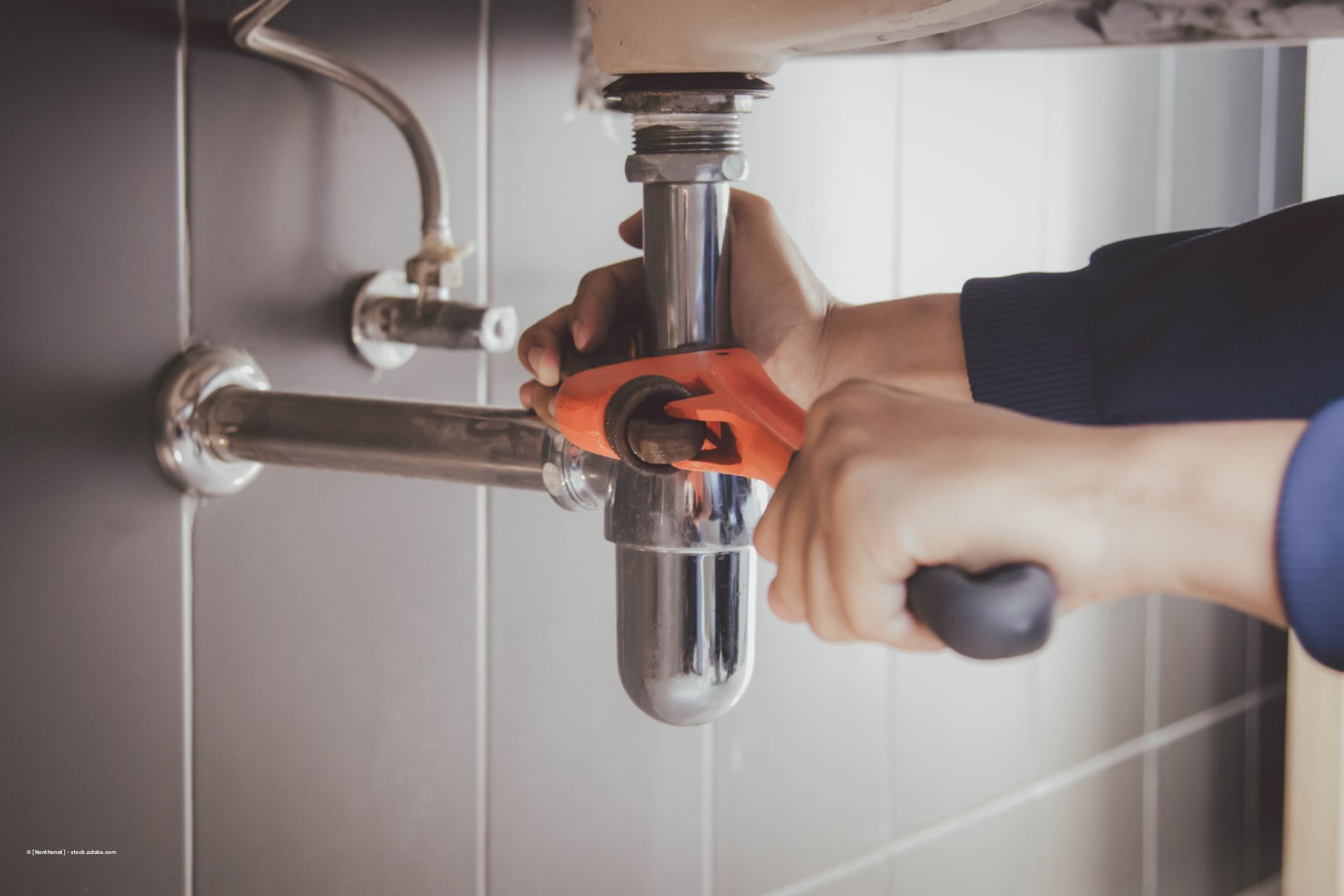 Person benutzt orangefarbenen Rohrschlüssel, um verchromten Siphon festzuziehen unter Waschbecken.