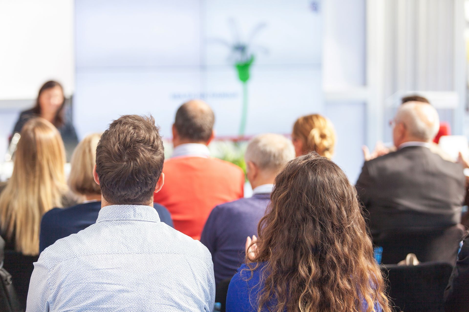 Le public est assis dans une salle de conférence lumineuse, face à un présentateur au premier rang.