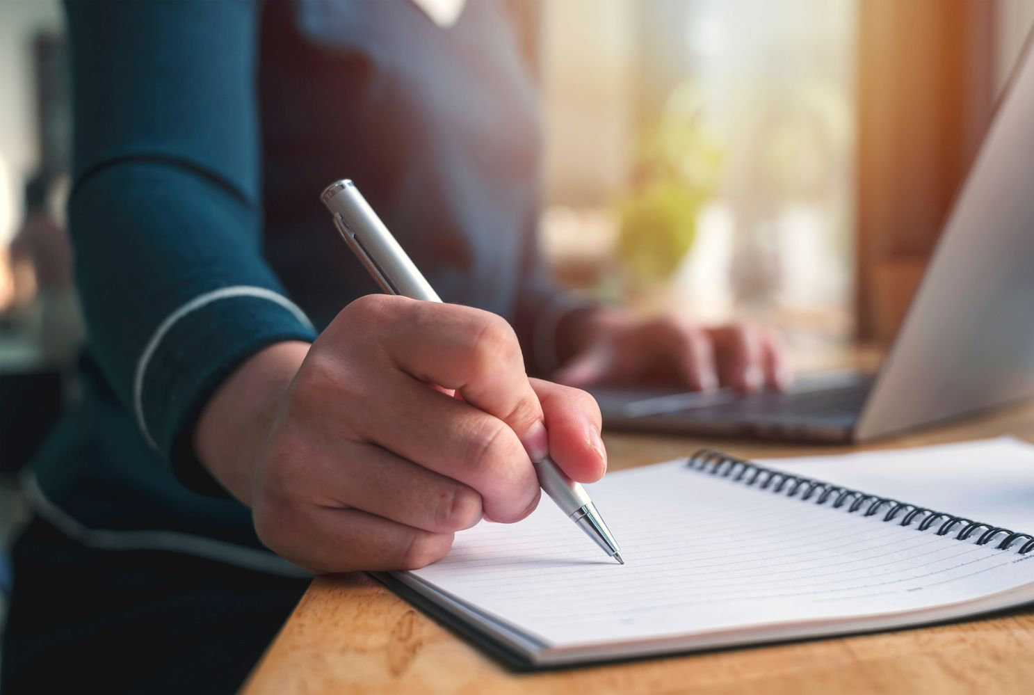 Une main tenant un stylo et écrivant dans un cahier à spirale sur un bureau ensoleillé