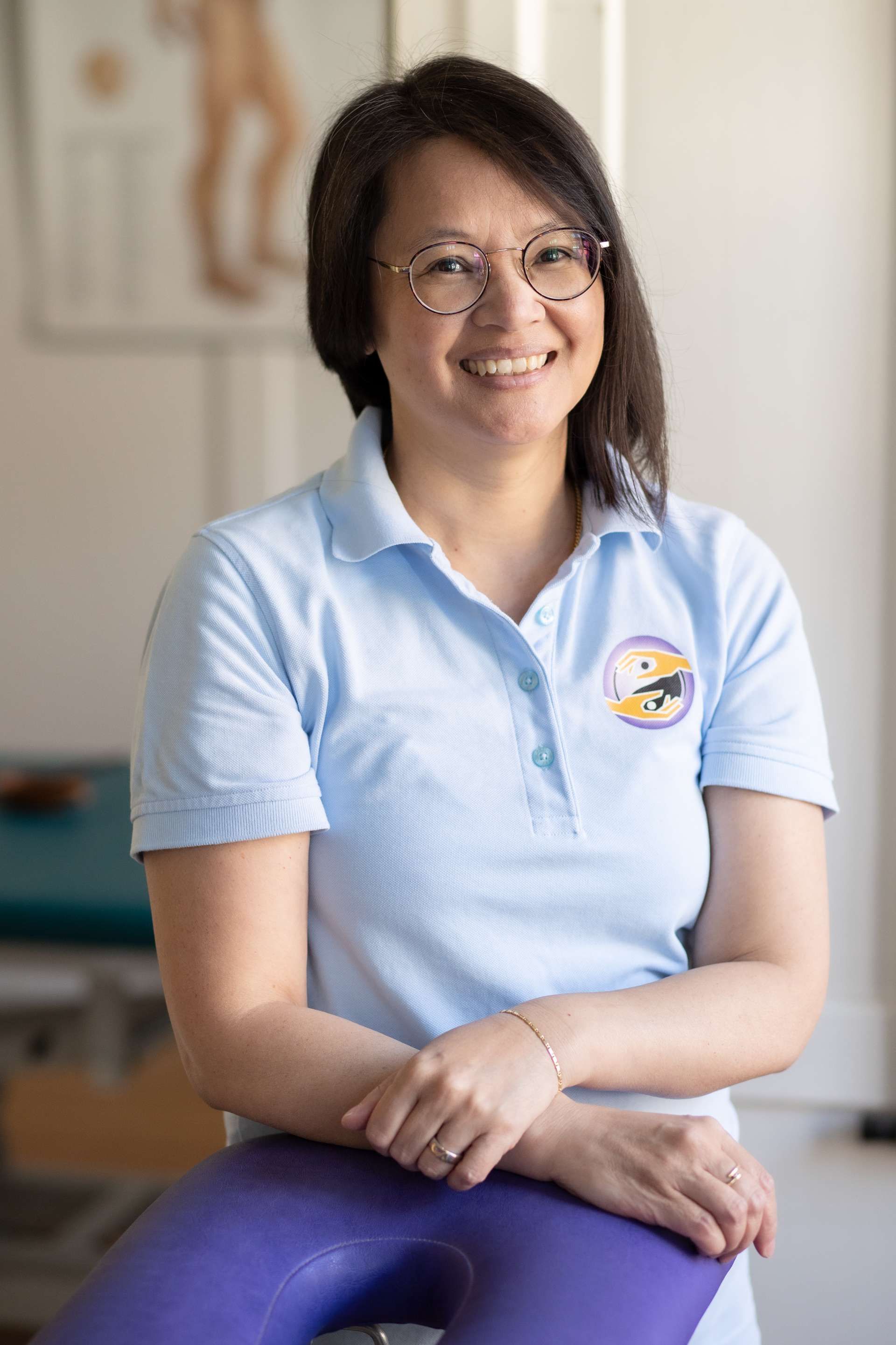 Frau mit Brille lächelt, trägt ein blaues Poloshirt und sitzt in der Nähe einer Massageliege in einem Innenraum.