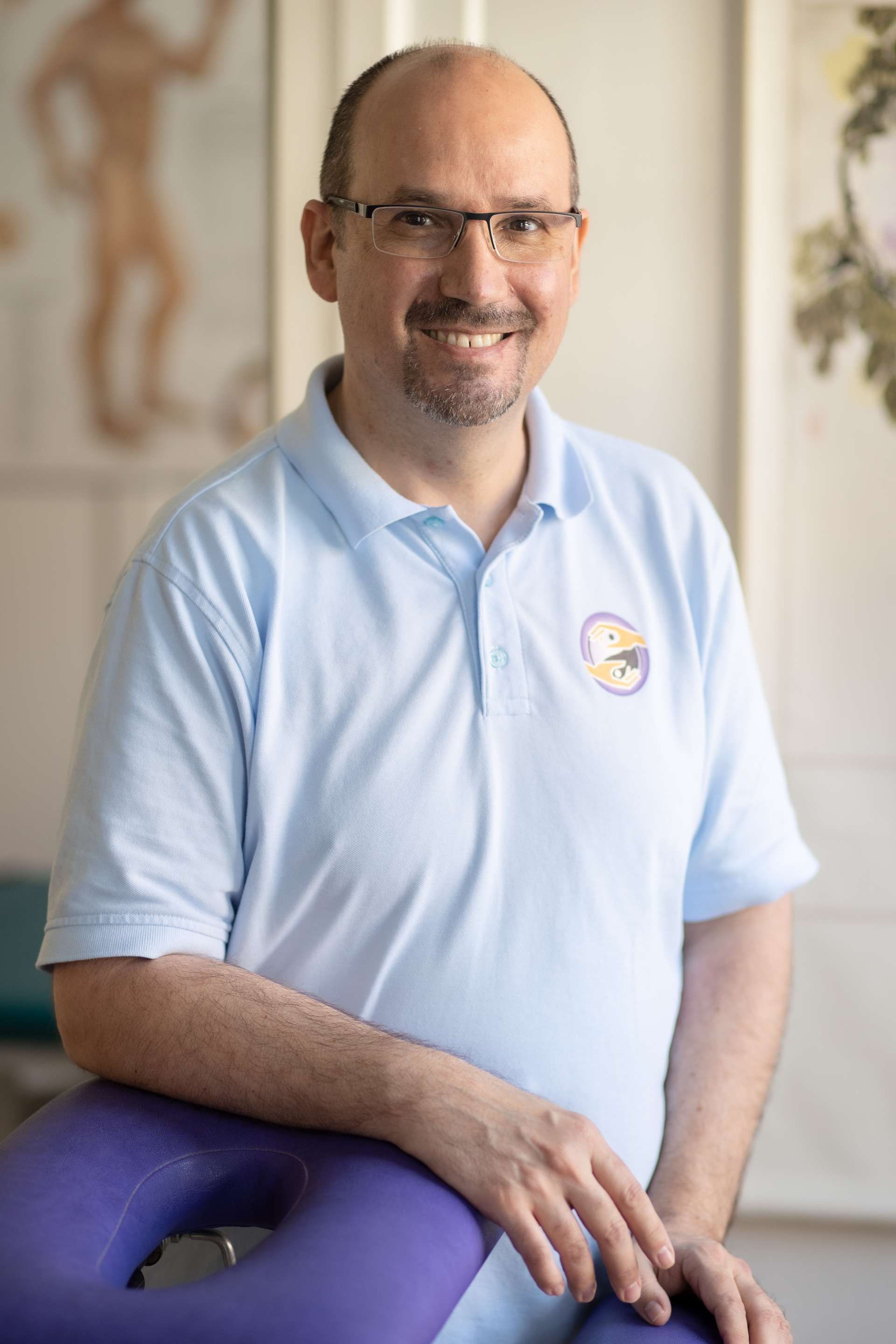 Ein Mann mit Brille und hellblauem Poloshirt lächelt und lehnt sich an eine violette Massageliege; Klinikatmosphäre.