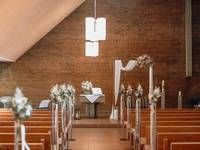 Ein modernes Kircheninterieur mit Reihen von Holzbänken, die auf einen mit weissen Blumen und einem Stoffbogen verzierten Backsteinaltar ausgerichtet sind.