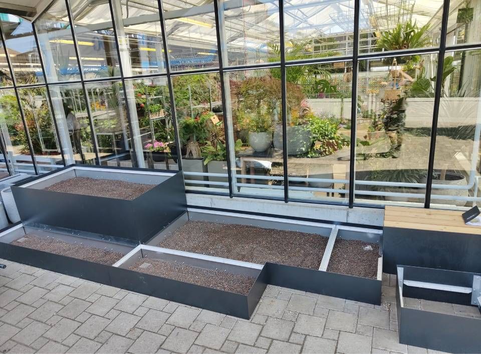 Graue, mehrstufige Metallpflanzkästen, gefüllt mit Erde, auf einer Terrasse innerhalb eines verglasten Gewächshauses.