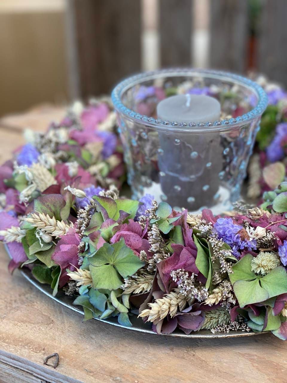 Ein gläserner Kerzenhalter mit einer grauen Kerze, umgeben von einem Kranz aus getrockneten Blumen in Lila-, Grün- und Beigetönen.
