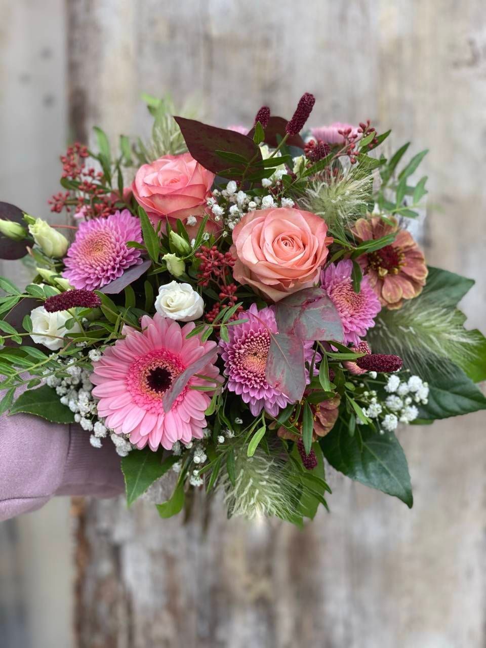 Ein handgehaltener Strauss aus rosa Rosen, Gerbera-Gänseblümchen und lila Nelken mit Schleierkraut und Grünzeug.