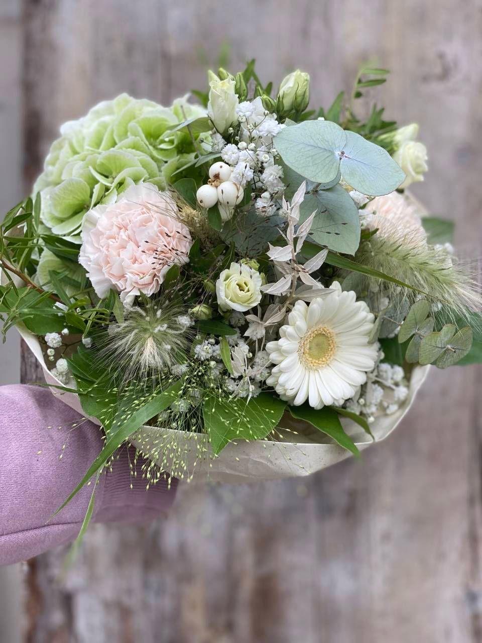 Ein Strauss aus hellrosa Nelken, weissen Gerbera-Gänseblümchen, weissen Beeren und grünen Hortensien, verpackt in Papier.