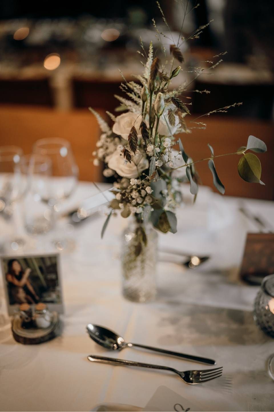 Ein florales Gesteck mit weissen Rosen und Grünzeug in einer Glasflasche auf einem gedeckten Esstisch mit Silberbesteck.