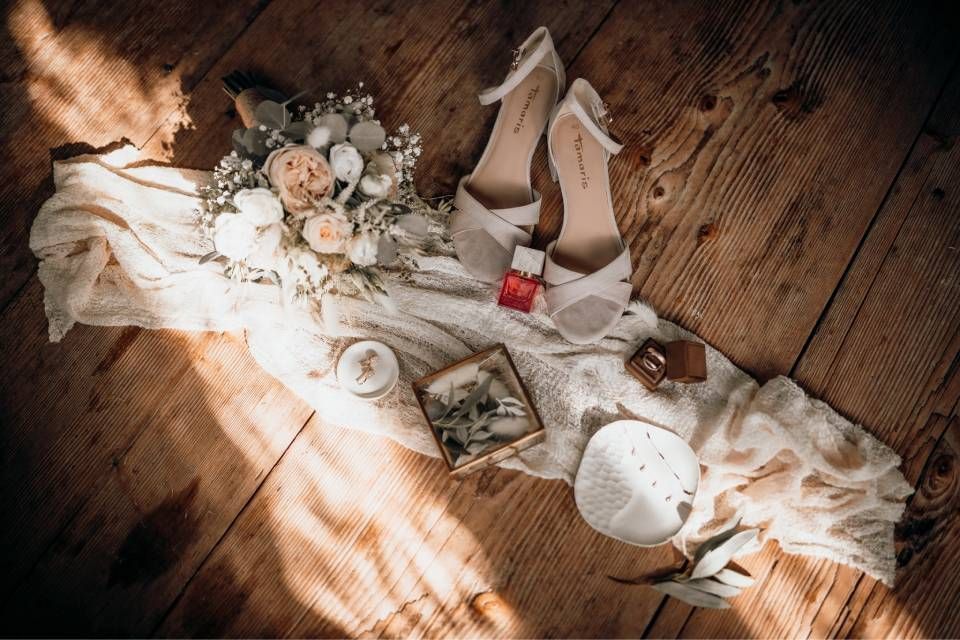 Hochzeitsaccessoires wie Schuhe, ein Brautstrauss, Ringe und dekorative Elemente sind auf einem weissen Läufer auf einem Holzboden arrangiert.