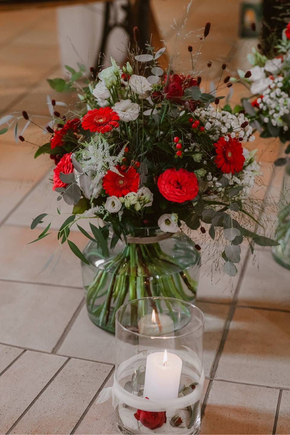 Ein Blumenarrangement mit roten und weissen Blumen in einer Glasvase, darunter eine brennende Kerze in einem Glashalter auf dem Boden.