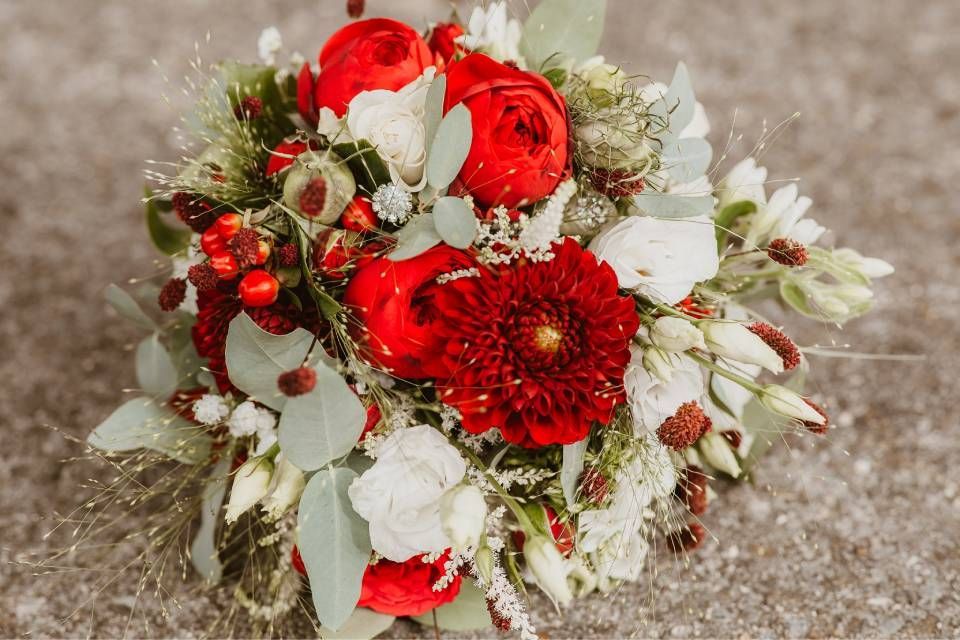 Ein Brautstrauss aus leuchtend roten Dahlien und Rosen, weissen Blüten und grünen Eukalyptusblättern auf Beton.