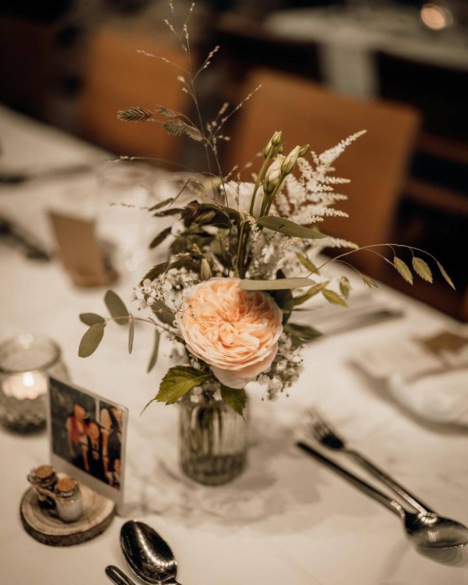 Ein florales Gesteck in einem Glasgefäss mit einer grossen pfirsichfarbenen Rose, Grünzeug und kleinen weissen Blüten auf einem Esstisch.