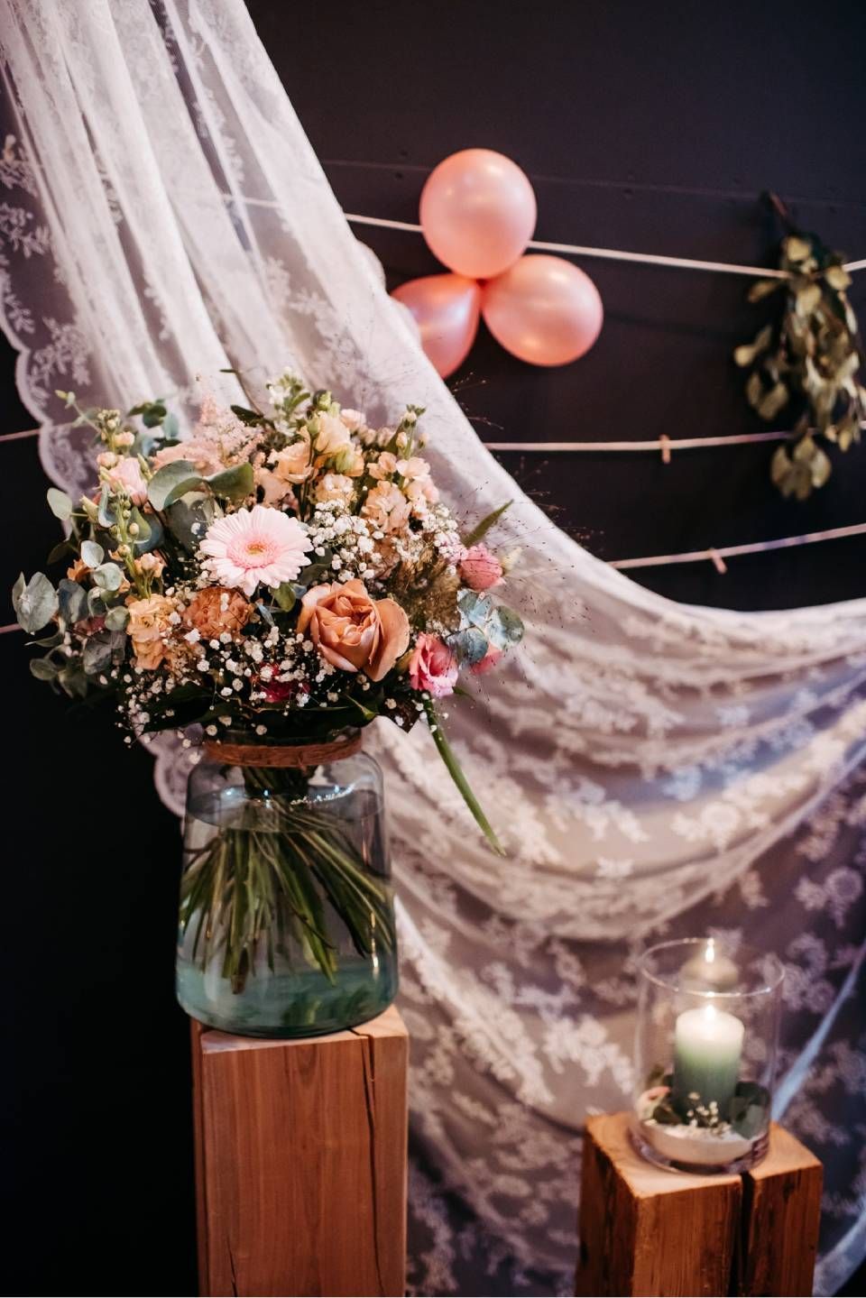 Ein Blumenstrauss in einer Glasvase und eine brennende Kerze stehen auf Holzsockeln vor einem Hintergrund aus Spitze und rosa Luftballons.