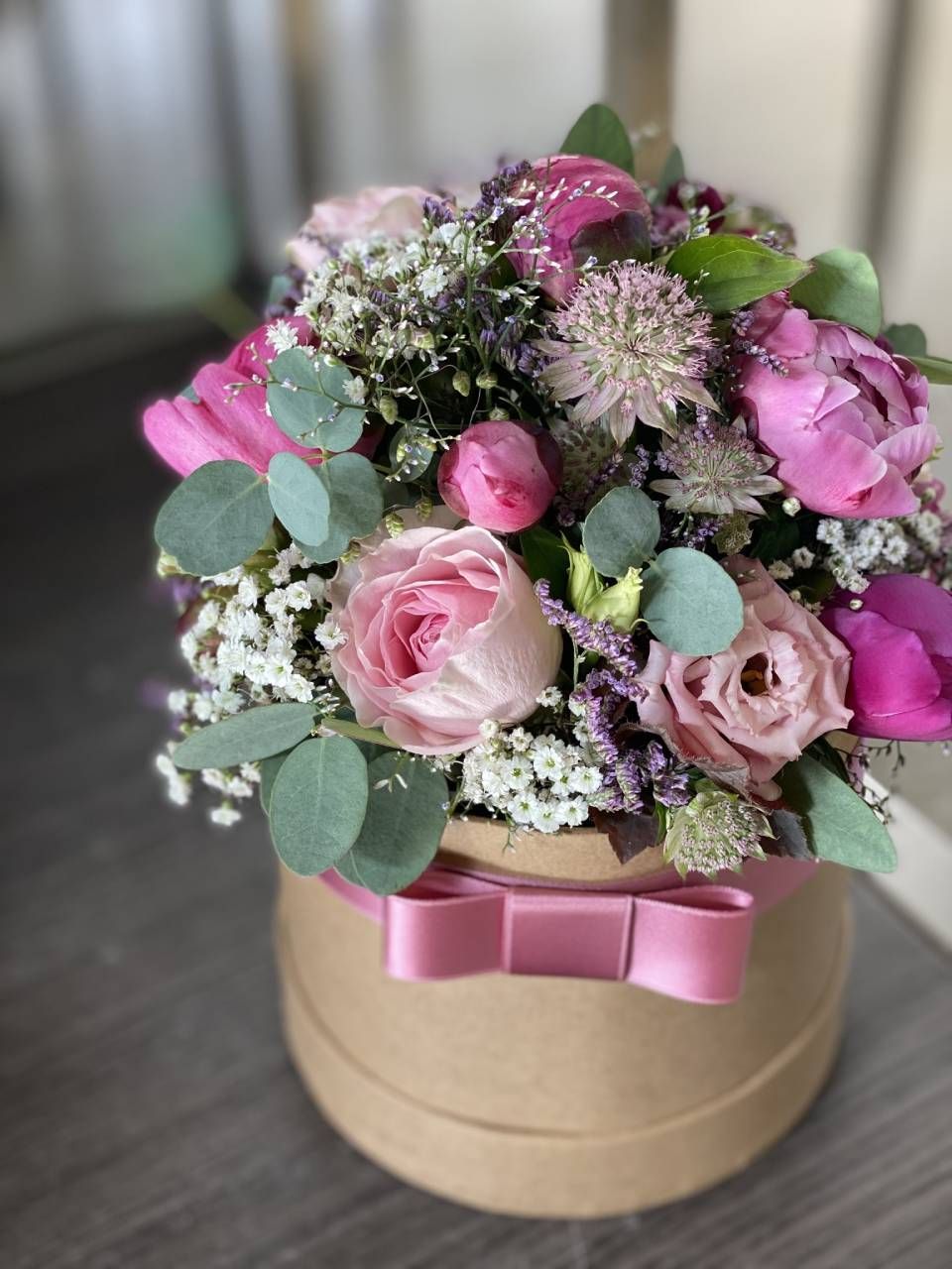 Eine runde, beige Geschenkbox mit rosa Schleife enthält einen Strauss aus rosa Rosen, Pfingstrosen und Grünzeug mit Schleierkraut.