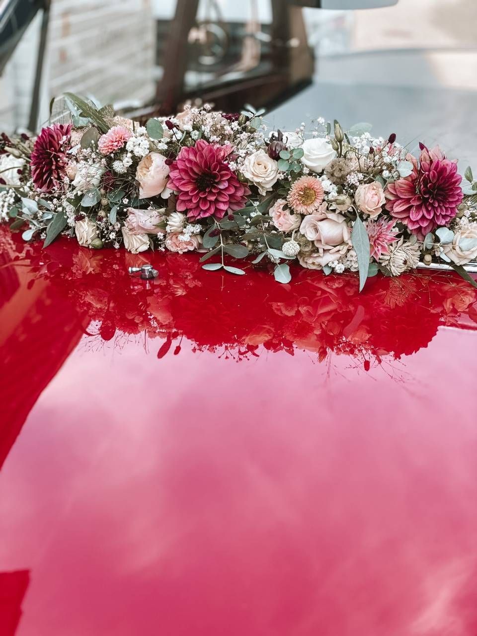 Ein grosses Blumenarrangement mit dunkelroten Dahlien, hellrosa Rosen und Grünzeug steht auf der Motorhaube eines roten Autos.