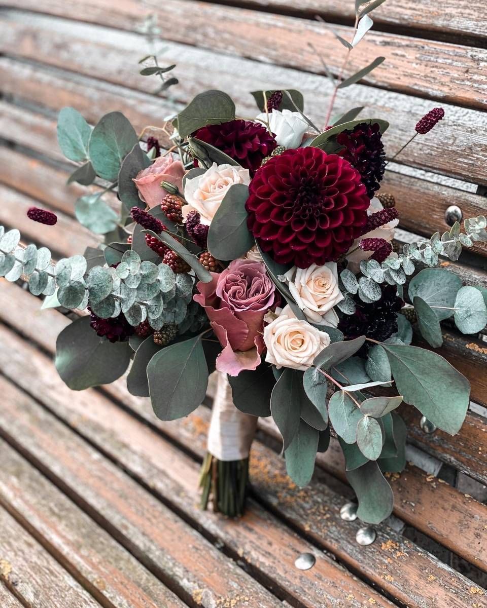 Ein üppiger Brautstrauss aus tief burgunderroten Dahlien, rosafarbenen Rosen und Eukalyptusblättern ruht auf einer Holzbank.
